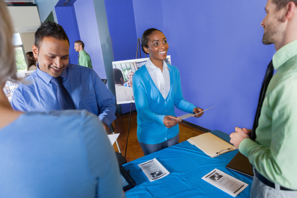 Man and woman welcome candidates to their booth at an IT job fair.
