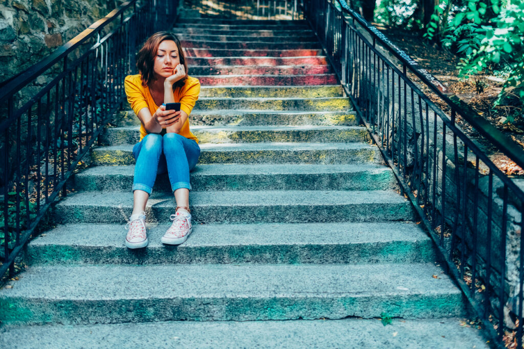 Woman sits on steps looking at her phone after being ghosted.